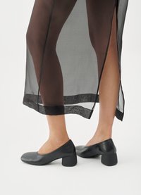 Black leather block-heel shoes with a rounded toe. Smooth, matte texture; low-cut design at the vamp. Worn with a sheer black skirt.