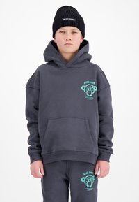 Jongen draagt een donkergrijze hoodie en bijpassende broek met turkooizen apengezichtlogo's, en een zwarte gebreide muts tegen een witte achtergrond.