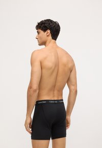 Boxer briefs de algodón negro con una cinturilla contrastante negra que presenta el logo "Calvin Klein", vistos desde la parte trasera en un modelo masculino.