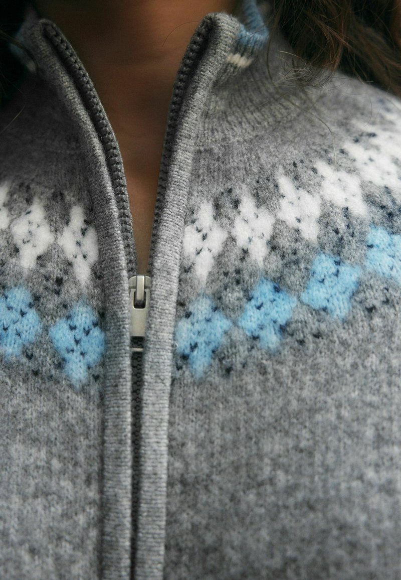 Gros plan sur un pull gris en tricot avec une fermeture éclair, présentant des motifs de losanges blancs et bleus près du col.