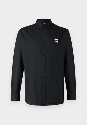 Musta pikka varrukaga nööbiga särk kraega, mille vasakul rinnal on väike valge joonistus näost prillides.