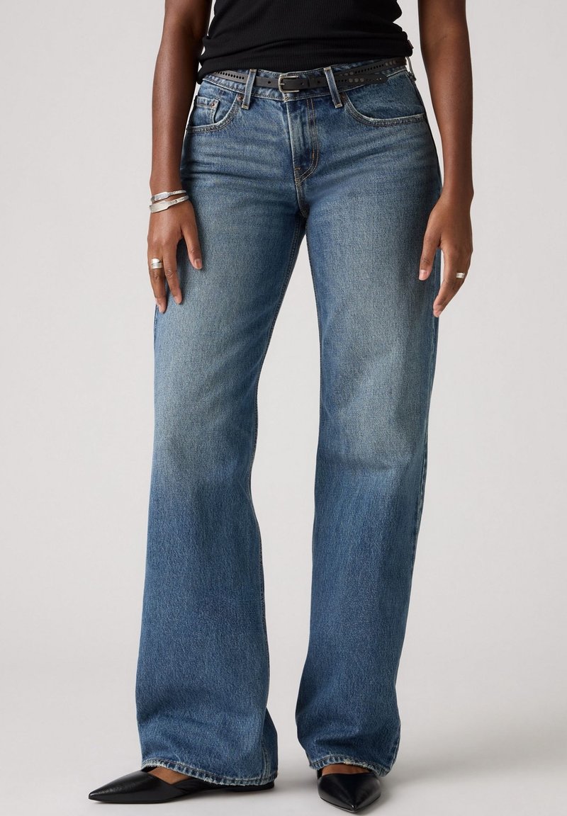 Personne portant un jean évasé bleu, une ceinture noire, des chaussures noires pointues, des bagues et bracelets en argent, debout devant un fond uni.