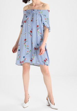 Frau trägt ein blau-weiß gestreiftes off-Shoulder-Kleid mit Blumenmuster und weiße Slingback-Pumps mit Blockabsatz, stehend vor weißem Hintergrund.