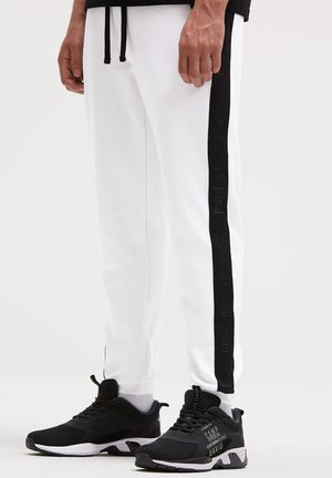 Lower body wearing white sweatpants with a black vertical stripe and black athletic shoes with white soles.