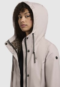 Veste imperméable beige avec capuche, cordon de serrage noir, doublure en imprimé léopard, boutons-pression et accent logo sur la poitrine.