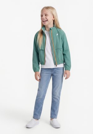 Mladé dievča sa smeje, má na sebe svetlozelenú bundu na zips, biele tričko, svetlomodré rifle a biele slip-on topánky, stojí na bielom pozadí.