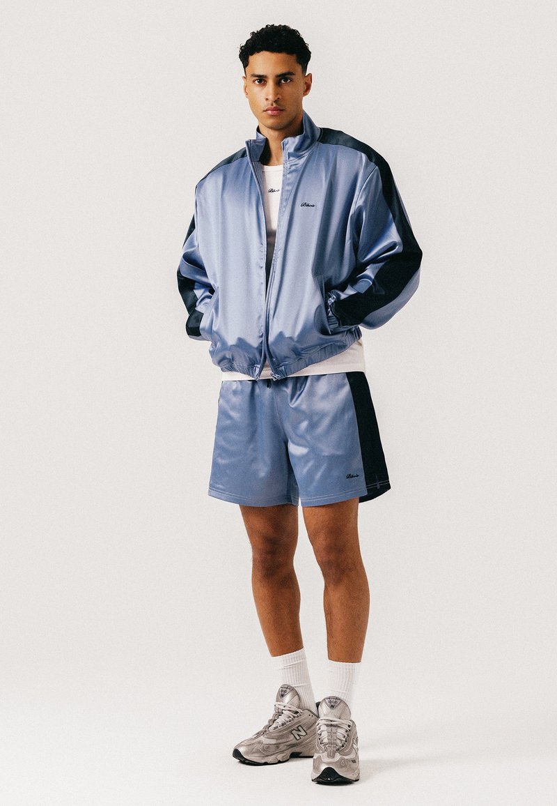 Jeune homme debout, les mains dans les poches, portant une veste de survêtement en satin bleu clair et un short avec des rayures noires, des chaussettes blanches et des baskets grises.