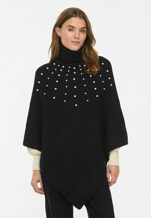 Femme portant un poncho noir à col roulé avec des ornements dispersés de perles blanches et des manches larges sur un haut à manches longues crème.