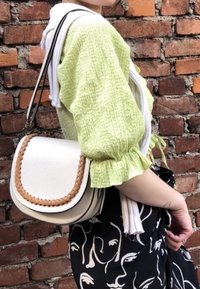 Witte leren zadelhandtas met gevlochten bruine accenten, crossbody gedragen. Model in een lichtgroene textuurblouse en een zwart met patroonrok.