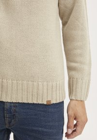 Beige neulottu villapaita, jossa on raidoitetut hihansuut ja helma, sekä pieni ruskea etiketti alareunassa. Yhdistetty sinisiin farkkuihin.