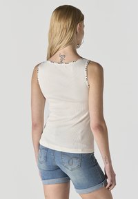 Ženska z dolgimi blond lasmi, oblečena v belo brezrokavnik in modre zavihane jeans kratke hlače, obrnjena stran na enobarvni podlagi.