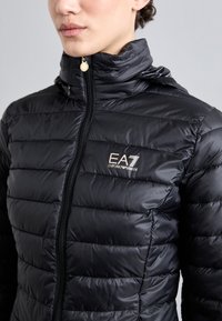 Črn puhovka z sijočo površino, ki ima vodoravno prešito zasnovo, stoječ ovratnik in zlat logo EA7 na prsih.