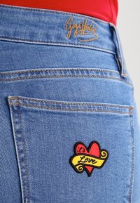 Ljusblå denimjeans med en gul logotyp på midjebandet och en hjärtapplikation på bakfickan, i röd och gul färg.