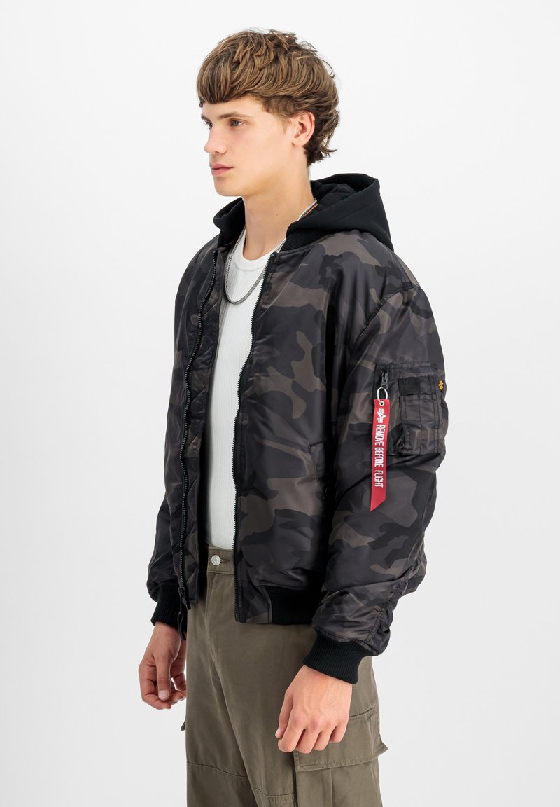 Giacca bomber camouflage con cappuccio nero, tasche con zip, polsini elasticizzati e un'etichetta rossa appesa alla manica. Tessuto dalla consistenza liscia.