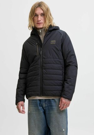 Chaqueta acolchada negra con cierre frontal, capucha y bolsillo en el pecho. Presenta una textura suave y un pequeño parche con el logotipo en el lado izquierdo.