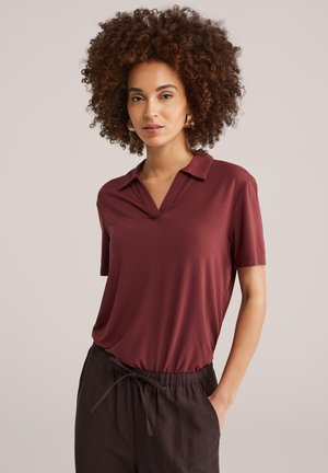 Käärus juustega naine kannab kuldset rõngakujulist kõrvarõngast, lühikeste varrukatega burgundiavärvi polosärki ja tumedaid nööritavaid pükse, üks käsi taskus.