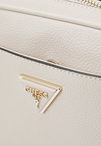 Cremefarvet læderhandtaske med tekstur, der har en triangel-formet guld- og hvid logo- accent på frontlommen, som har en lynlåslukning.