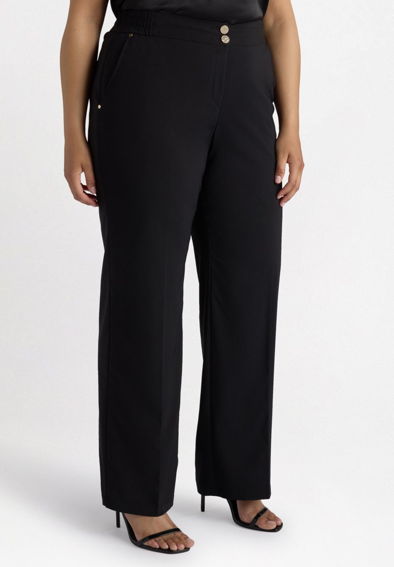 Svarta vida byxor med en slät textur, som har två metallknappar och sidofickor. Kombineras med svarta högklackade sandaler.