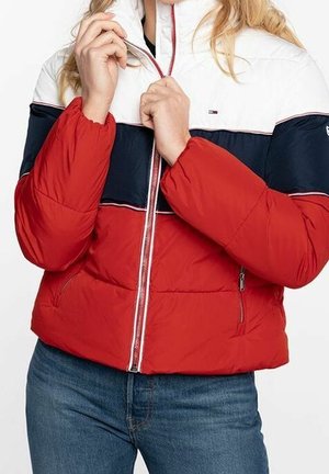Person, der bærer en rød, navy- og hvid foret jakke, og er ved at lynlåse den, med blå jeans synlige fra taljen og ned.