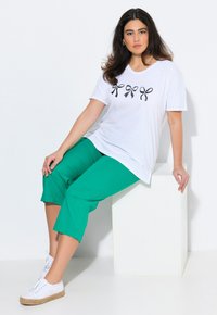 Weißes kurzes T-Shirt mit schwarzen Schleifenmuster, kombiniert mit grünen Caprihosen. Das Model trägt weiße Schnürschuhe und sitzt auf einem weißen Würfel.