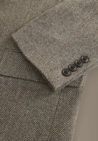 Manche de blazer en laine à motif chevrons marron et beige avec quatre boutons marron foncé sur le poignet.