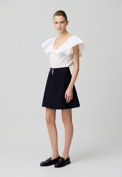 Blouse blanche avec des manches à volants volumineuses, détails en dentelle, associée à une jupe noire évasée et des mocassins noirs avec une touche dorée.