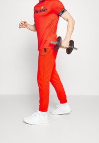 Röd träningsoutfit bestående av en t-shirt med kort ärm och joggingsbyxor, kombinerad med vita sneakers, hållande en hantel i ena handen.