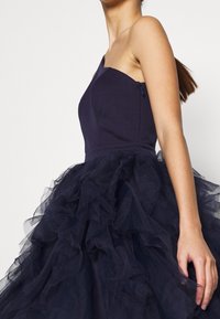 Laivastonsininen mekko, jossa istuva vartalo ja yksittäinen olkapääntalja, tilava tyllihame, jossa on röyhelöitä ja sivuvetoketju.