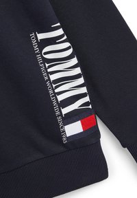 Granatowa bluza z wyraźnym białym napisem „TOMMY” i detalami marki. Posiada czerwone, białe i niebieskie paski na dole.