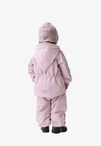 Lichtroze waterdichte jas met een capuchon, een elastische taille, gecombineerd met bijpassende broek. De outfit wordt aangevuld met een beige gebreide muts.