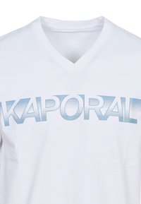Camiseta blanca de cuello en V hecha de algodón con un estampado gráfico en azul claro del nombre de la marca "KAPORAL" que presenta detalles punteados.