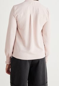 Chemise rose clair à manches longues avec une finition texturée et des poignets smockés, associée à un pantalon sombre à fines rayures.