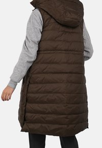 Person wearing long, dark brown padded vest with hood over a gray long-sleeve shirt, shown from the back and mid-thigh down.