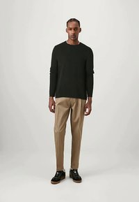 Férfi áll, fekete hosszúujjú pulóverben, bézs nadrágban és fekete, barna talpú sportcipőben egy sima világos háttér előtt.