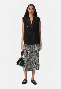 Czarny dwurzędowy bezrękawnik, plisowana spódnica w czarno-białe wzory, czarna torba z ozdobnymi uchwytami oraz czarne płaskie buty.