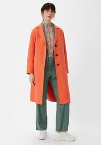 Oranger Wollmantel mit Reverskragen und vier schwarzen Knöpfen, getragen über einer gemusterten Bluse und türkisfarbenen Hosen. Weiße Sneakers vervollständigen das Outfit.