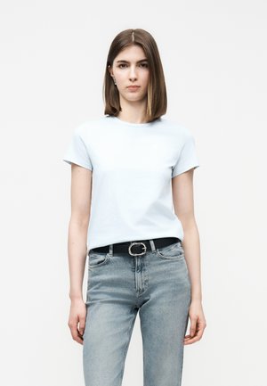 Mladá žena s rovnými hnedými vlasmi, oblečená v svetlomodrom tričku s krátkym rukávom a svetlomodrých džínsoch s čiernym opaskom, stojaca na bielom pozadí.