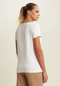 Camiseta blanca de manga corta con escote redondo, hecha de tela suave. Presenta un pequeño logo rosa en la parte posterior del cuello y un ajuste relajado.