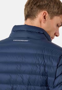 Giacca imbottita blu navy con texture trapuntata e colletto alto. Presenta un logo SWEDEMOUNT ben visibile sul retro. Materiale liscio e leggero.