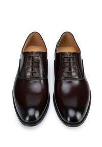 Zapatos de vestir de cuero marrón con acabado suave, punta redondeada y detalle de cordones en el centro. Los acentos cosidos añaden textura y refinamiento.