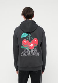 Tumši pelēks hoody ar ķiršu grafiku un tekstu "ADIDAS ORIGINALS". Materiāls izskatās mīksts, ar brīvu piegriezumu un ribotiem piedurkņu galiem.