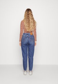 Blauwe high-waisted jeans met vijf zakken, een bruine leren patch op de achterkant en gefranjerde zomen, gecombineerd met een gebloemde lange mouwtop.