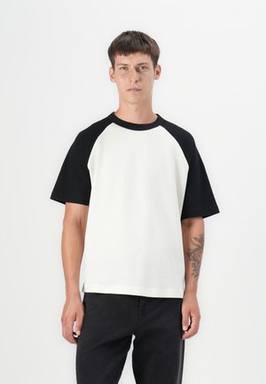 T-shirt raglan avec un corps blanc et des manches noires ; fabriqué en tissu texturé avec un col rond et des ourlets fendus sur les côtés.