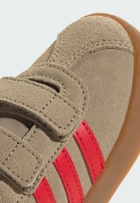 Bež nubuk cipela s crvenim kožnim trakama, s dva čičak zatvarača i gumiranim potplatom; vidljiv je detalj na gornjem dijelu.