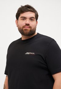 Tricou negru din bumbac, cu guler rotund. Are un logo mic în alb și roșu pe piept, cu mâneci scurte și o textură netedă.