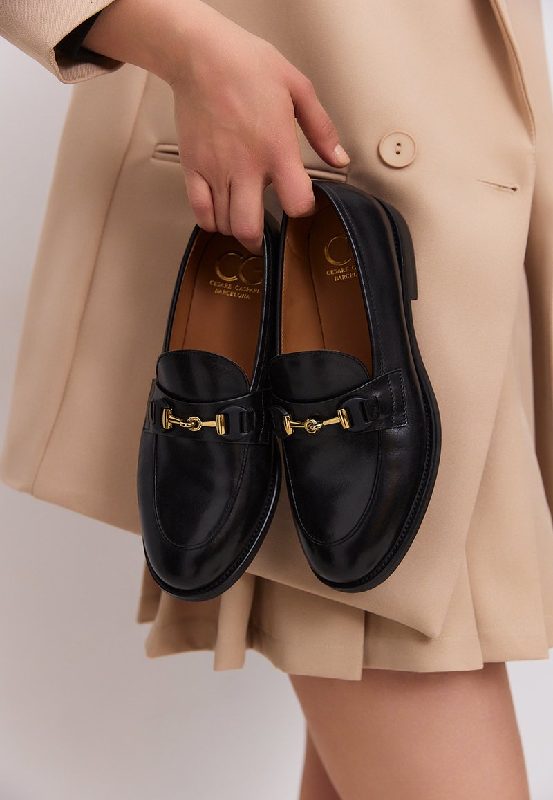 Zwarte leren loafers met een ronde neus, decoratieve gouden hardware en een gladde afwerking, vastgehouden door een hand tegen een beige outfit achtergrond.