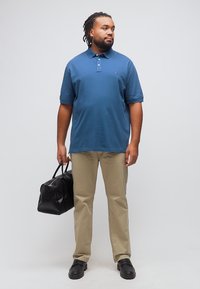 Άνδρας με πλεγμένα μαλλιά, φορά μπλε πόλο, μπεζ παντελόνι, μαύρα παπούτσια, κρατώντας μαύρη υφασμάτινη τσάντα duffel, κοιτάζει προς το πλάι σε απλό φόντο.