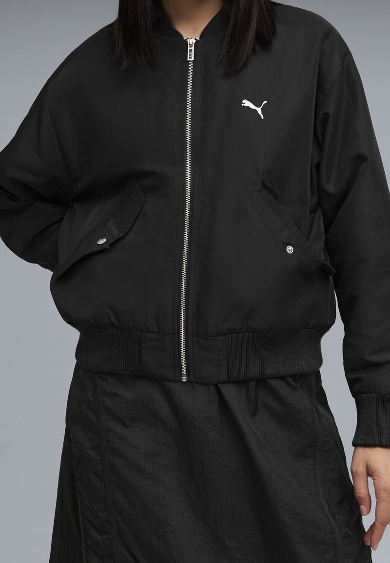 Personne portant une veste Puma noire zippée avec poignets et taille côtelés, poches avant avec boutons, et une jupe noire texturée.