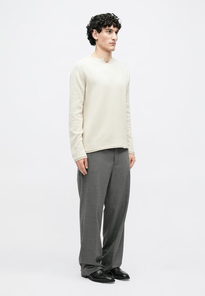 Ανοιχτό μπεζ πλεκτό πουλόβερ με στρογγυλή λαιμόκοψη, συνδυασμένο με γκρι παντελόνι φαρδύ στο πόδι. Μαύρα παπούτσια ολοκληρώνουν το ντύσιμο. Απλός σχεδιασμός και ελάχιστες λεπτομέρειες.