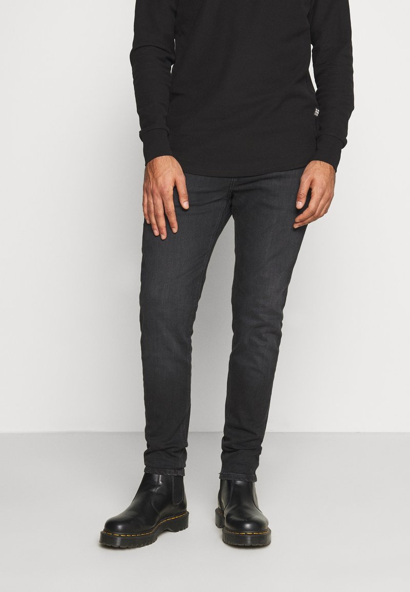 Camisa negra de manga larga combinada con jeans de denim oscuros, con un corte ajustado. Botas de cuero negras con suela gruesa completan el conjunto.